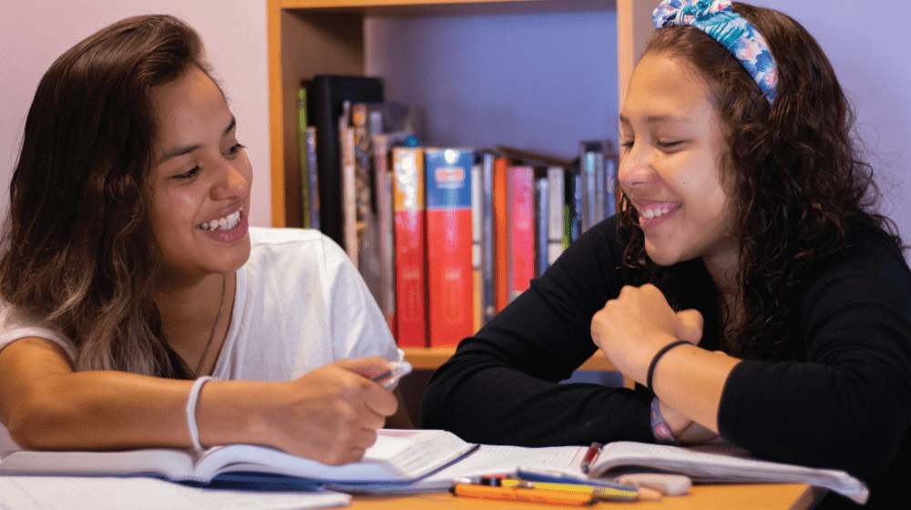 Dos mujeres adolescentes con un cuaderno abierto y un librero de fondo